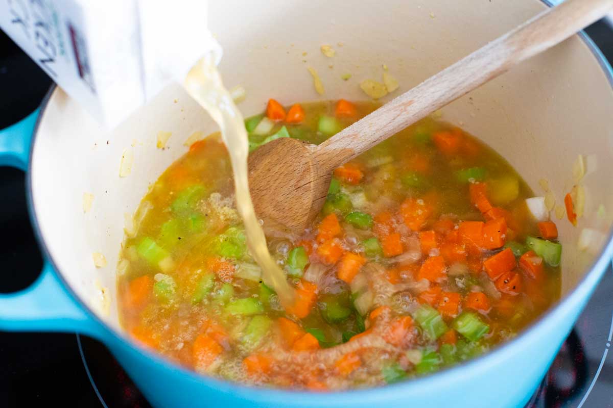 A soup post has chopped carrots and celery being stirred with a spoon while the other hand pours in chicken broth.