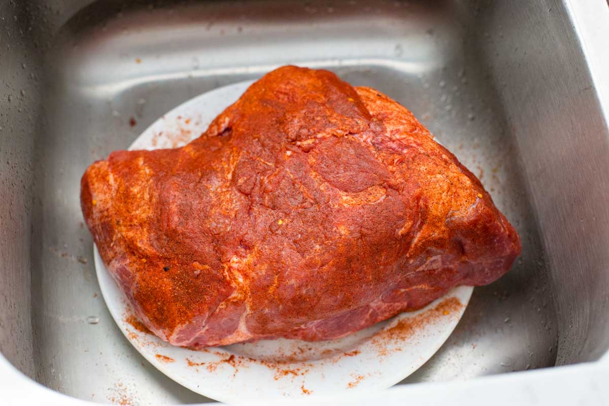 The pork shoulder sits on a clean plate in the sink and has been rubbed with spices all over.