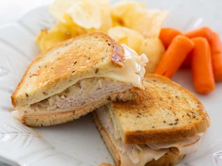 A grilled turkey reuben sandwich is on a plate with potato chips and baby carrots.