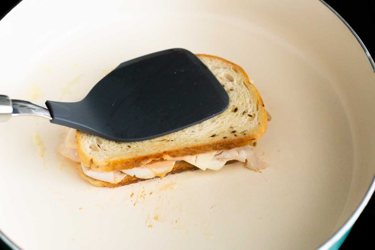 A flat spatula is pressing the sandwich as it griddles in the frying pan.