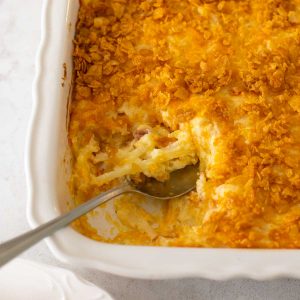 The funeral potatoes recipe has been baked and a spoon has scooped out a serving from the baker.