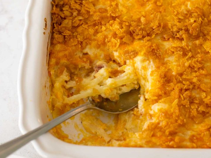 The funeral potatoes recipe has been baked and a spoon has scooped out a serving from the baker.