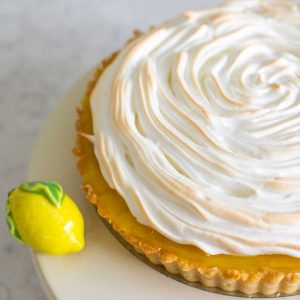 The finished lemon meringue tart is on a cake plate with a ceramic lemon decoration next to it.