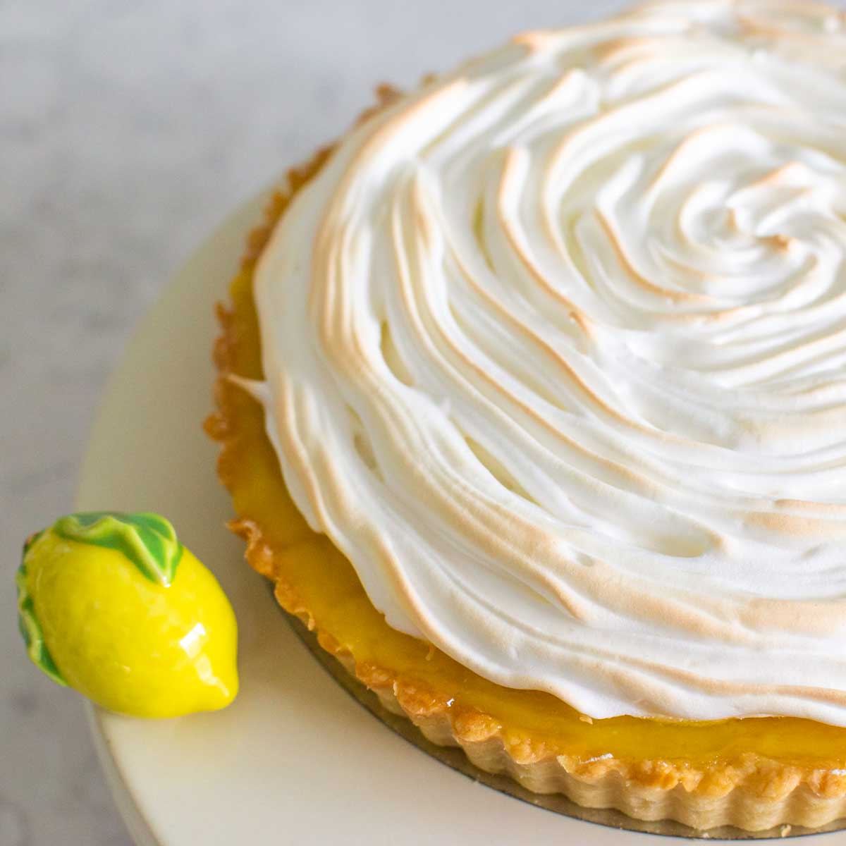 The finished lemon meringue tart is on a cake plate with a ceramic lemon decoration next to it.