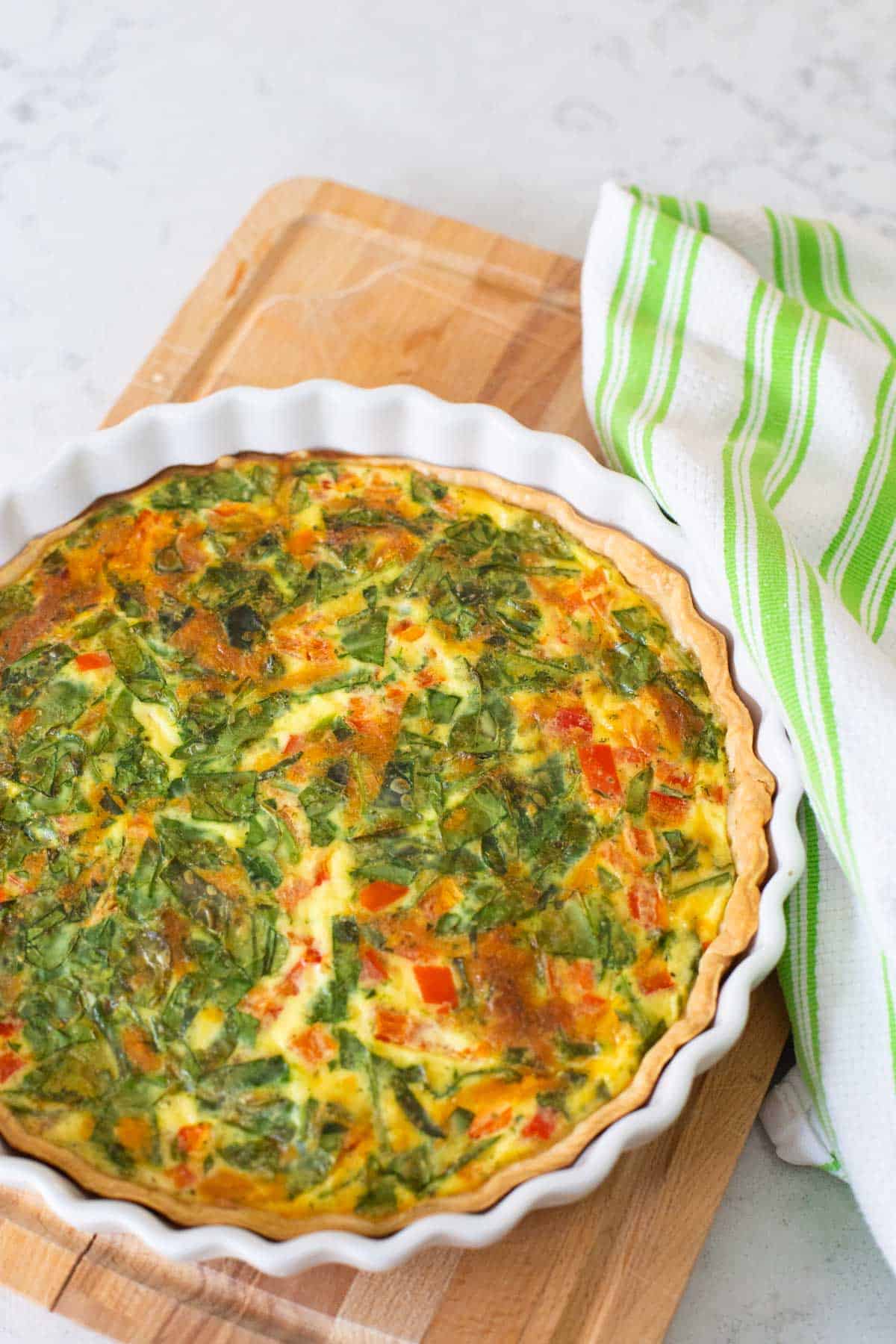 The quiche is cooling on a wooden cutting board with a towel next to it.