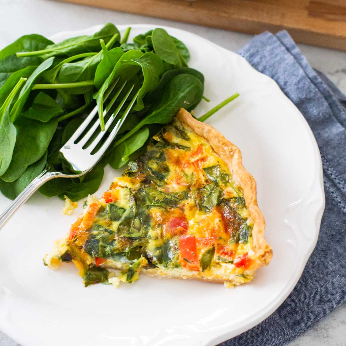 A slice of spinach quiche is on a plate next to additional baby spinach.