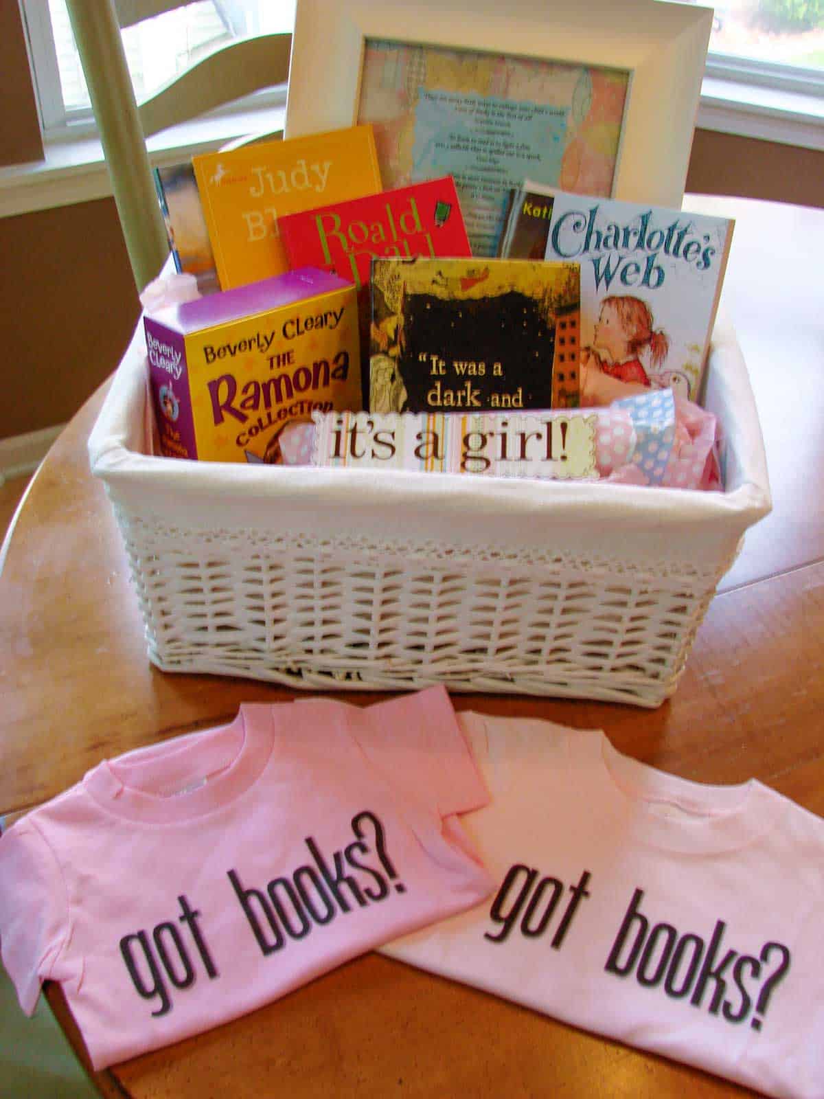 A basket of books to start a young girl's home library. There is a framed printable of book quotes in the basket and two pink onesies sitting on the table in front.