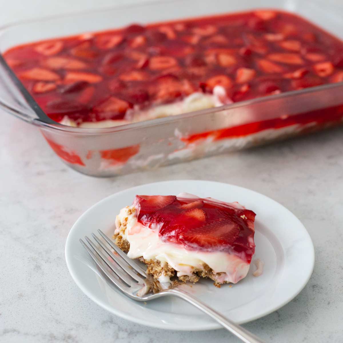 A 9x13-inch pan has finished strawberry pretzel salad for serving, a plate with a single slice and a fork is in front.