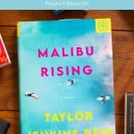 A copy of Malibu Rising sits on a table surrounded by books.