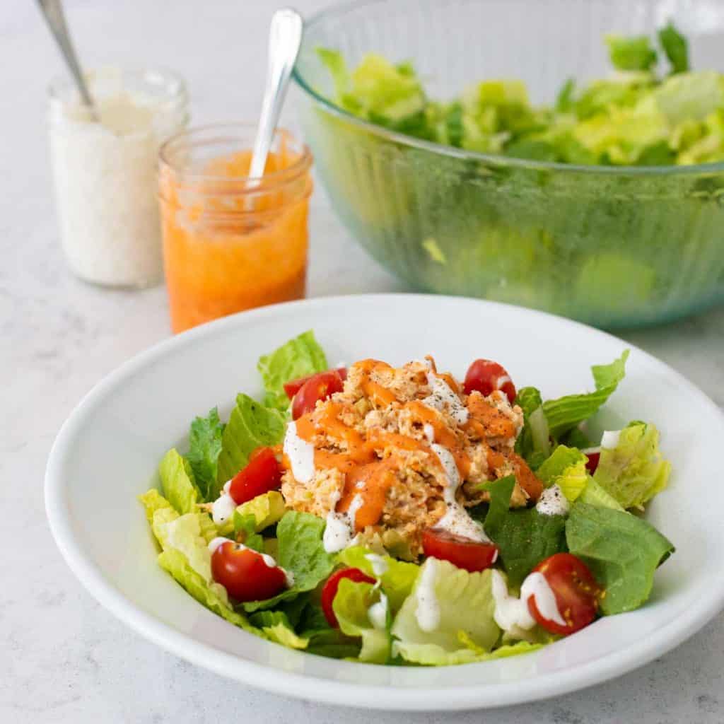 A bowl filled with green lettuce, topped with buffalo chicken salad, fresh tomatoes, and drizzled with buffalo sauce and blue cheese dressing sits in front of a serving bowl of lettuce with mason jars of dressing on the side.