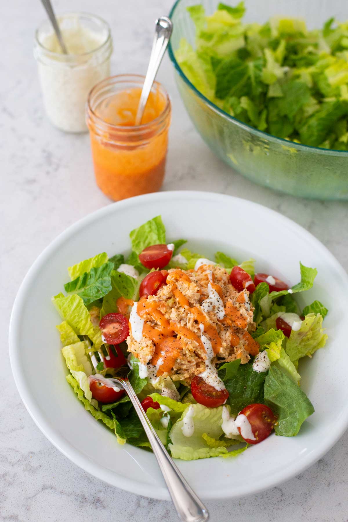 A serving of buffalo chicken salad sits in a white bowl in front of a bowl of lettuce with dressings on the side.