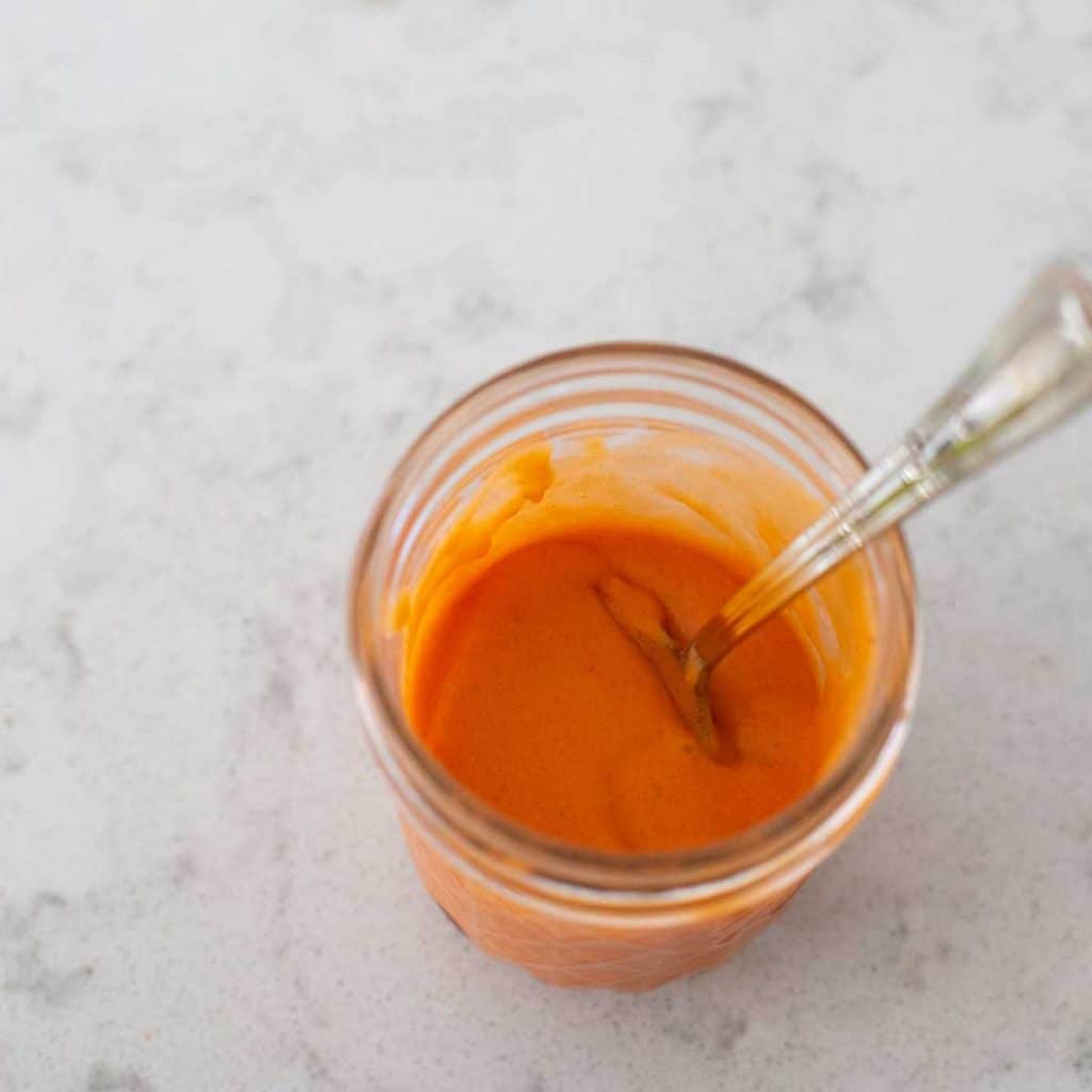 A super easy homemade buffalo sauce sits in a mason jar with a spoon.