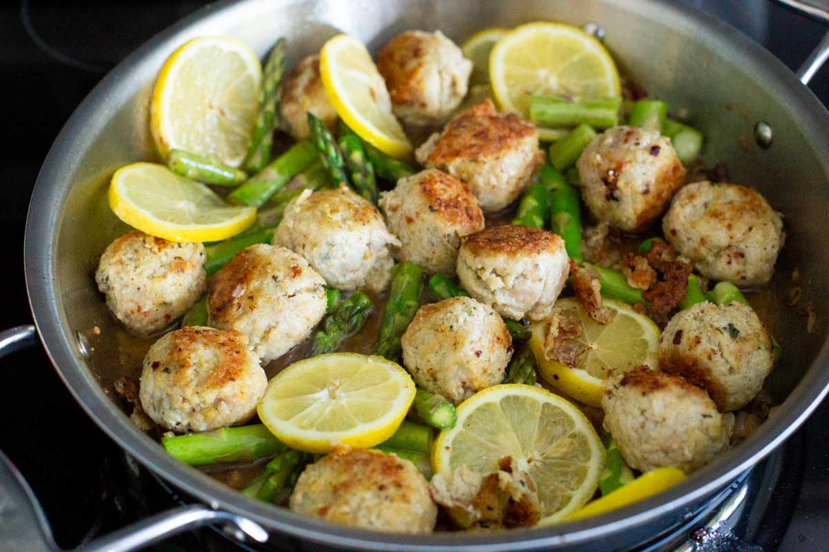 The skillet has the browned meatballs, fresh lemon slices, and asparagus.