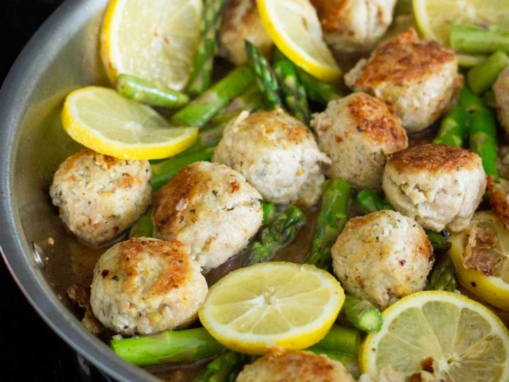 A skillet filled with meatballs, lemon slices, and asparagus spears.
