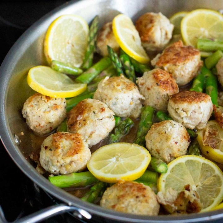 A skillet filled with meatballs, lemon slices, and asparagus spears.