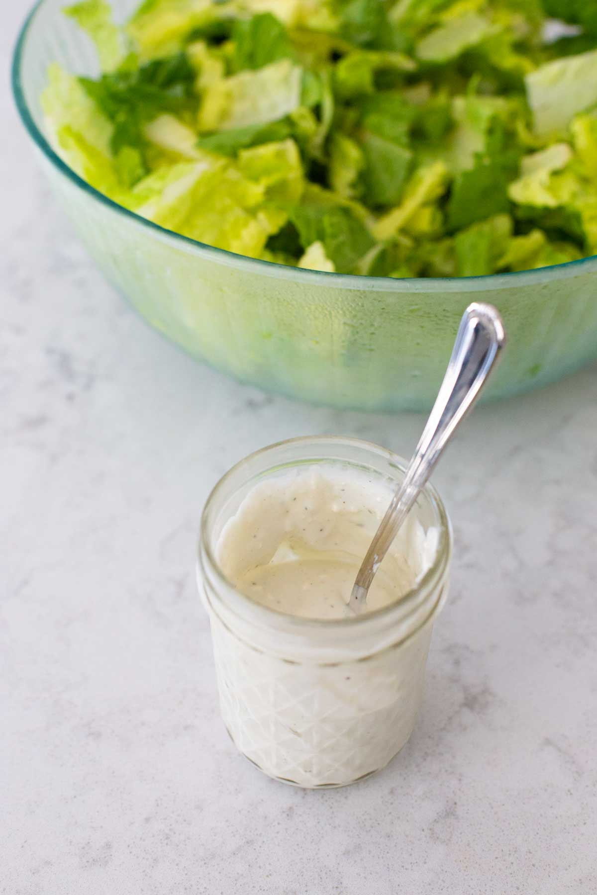 A mason jar filled with homemade blue cheese dressing has a spoon for serving and sits in front of a bowl of lettuce.