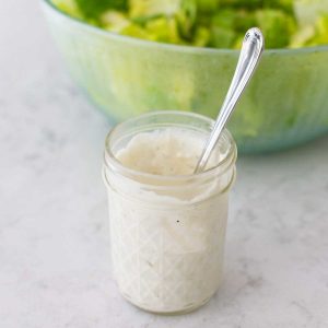 A mason jar filled with homemade blue cheese dressing sits in front of a bowl full of lettuce.