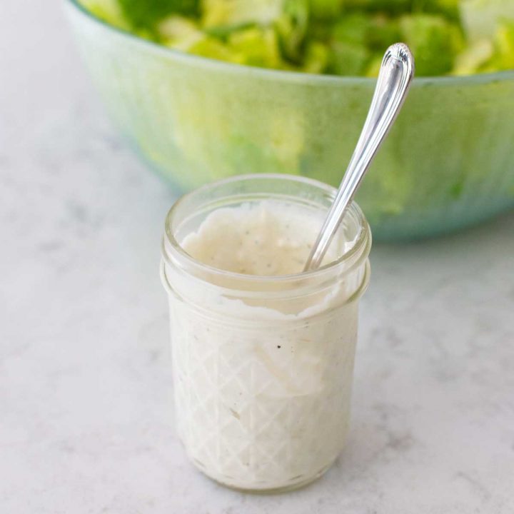 A mason jar filled with homemade blue cheese dressing sits in front of a bowl full of lettuce.