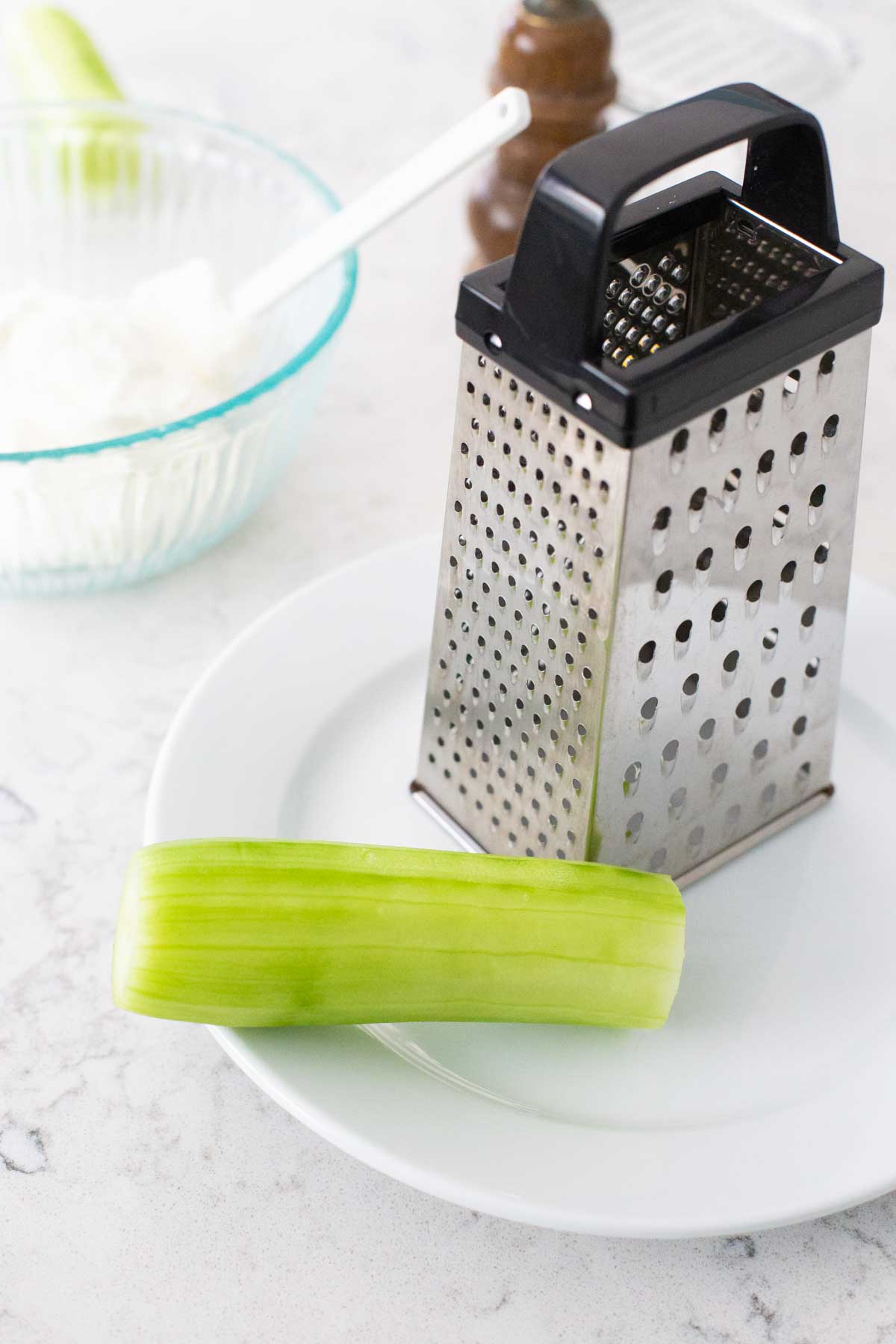 A fresh cucumber has been peeled and sits next to a box grater.