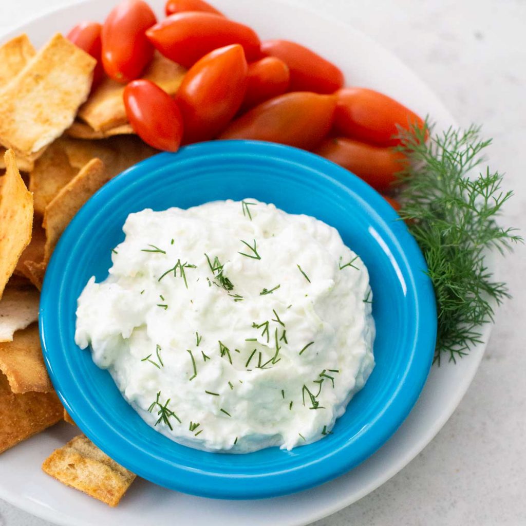 A blue bowl filled with homemade tzatziki sauce has fresh cherry tomatoes, pita chips for dipping, and a sprig of fresh dill.