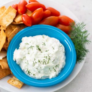 A blue bowl filled with homemade tzatziki sauce has fresh cherry tomatoes, pita chips for dipping, and a sprig of fresh dill.