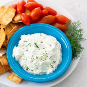 A blue bowl filled with homemade tzatziki sauce has fresh cherry tomatoes, pita chips for dipping, and a sprig of fresh dill.
