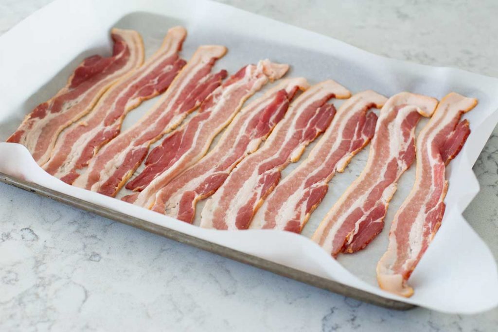 A baking sheet has been lined with parchment and the bacon is lined up in a single even layer.
