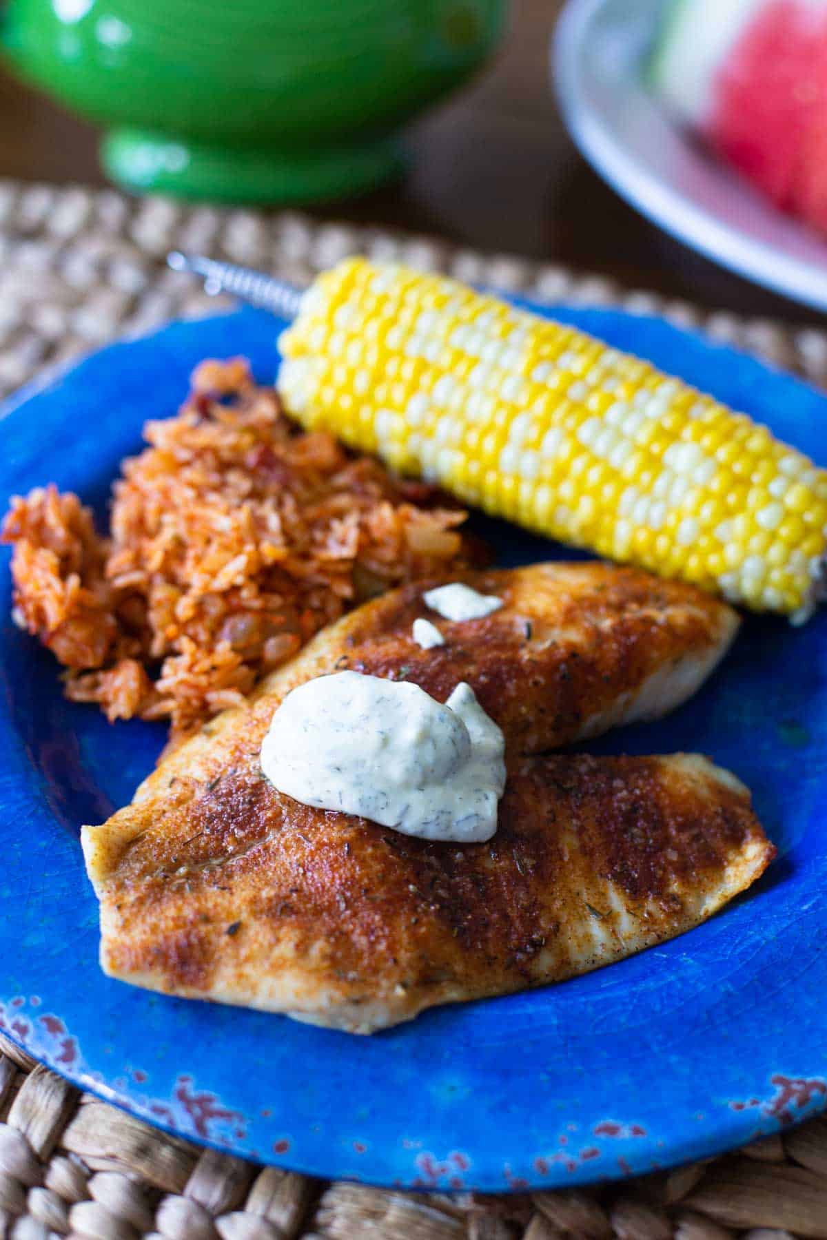 The grilled tilapia on a blue plate with a scoop of red rice and a cob of corn.