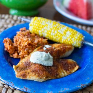 The grilled tilapia is on a dinner plate with a dollop of dill aoli, red rice, and a cob of corn.