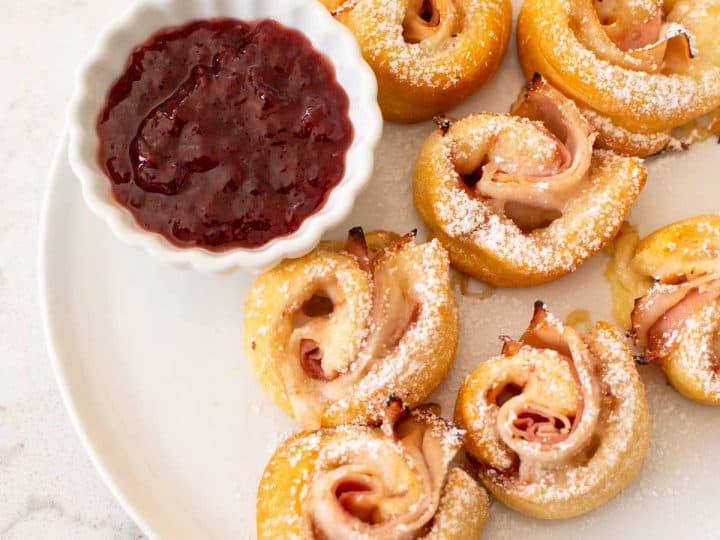 A plate filled with Monte Cristo crescent rolls and a little cup of strawberry jam for serving.