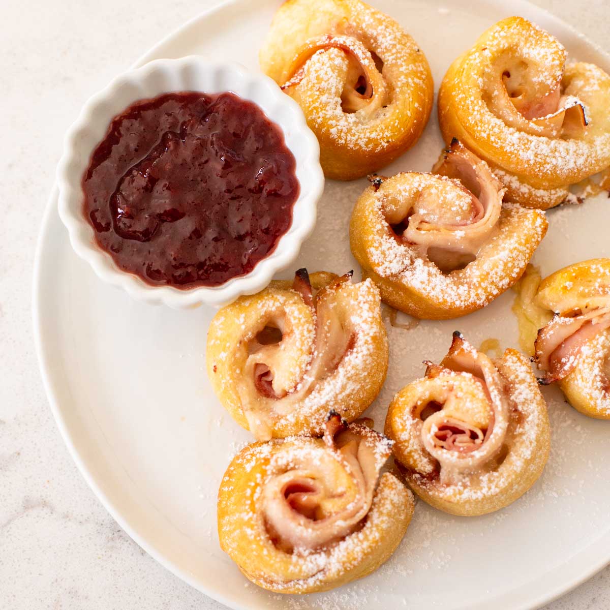 A plate filled with Monte Cristo crescent rolls and a little cup of strawberry jam for serving.