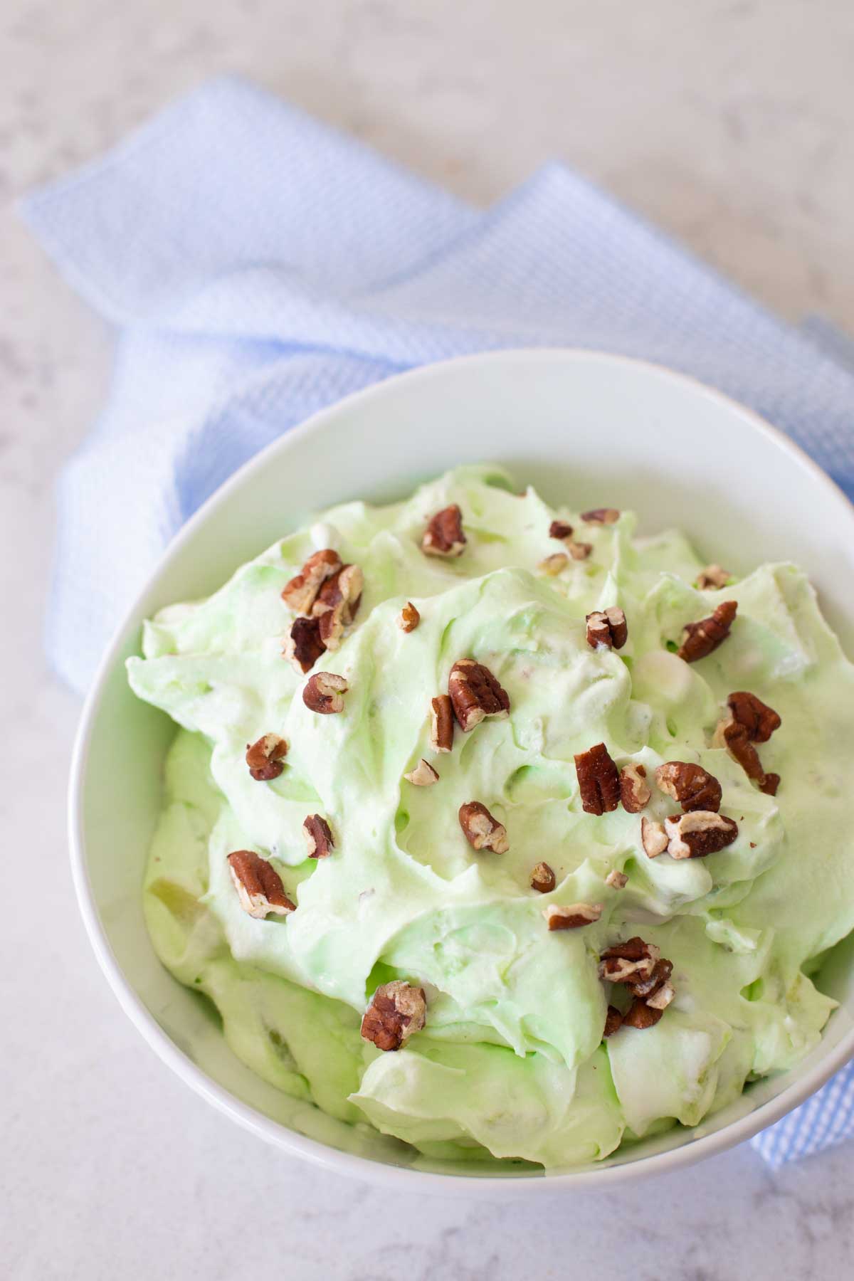 A serving bowl of pistachio fluff with chopped pecans sprinkled over the top has a blue napkin on the side.