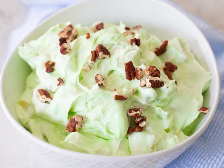 A large serving bowl of pistachio fluff has chopped pecans sprinkled over the top and a blue napkin on the side.