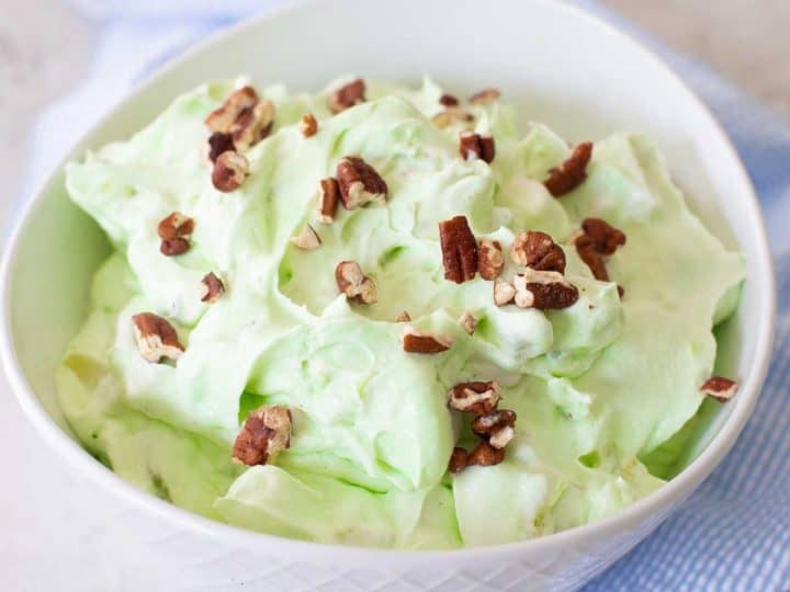 A large serving bowl of pistachio fluff has chopped pecans sprinkled over the top and a blue napkin on the side.