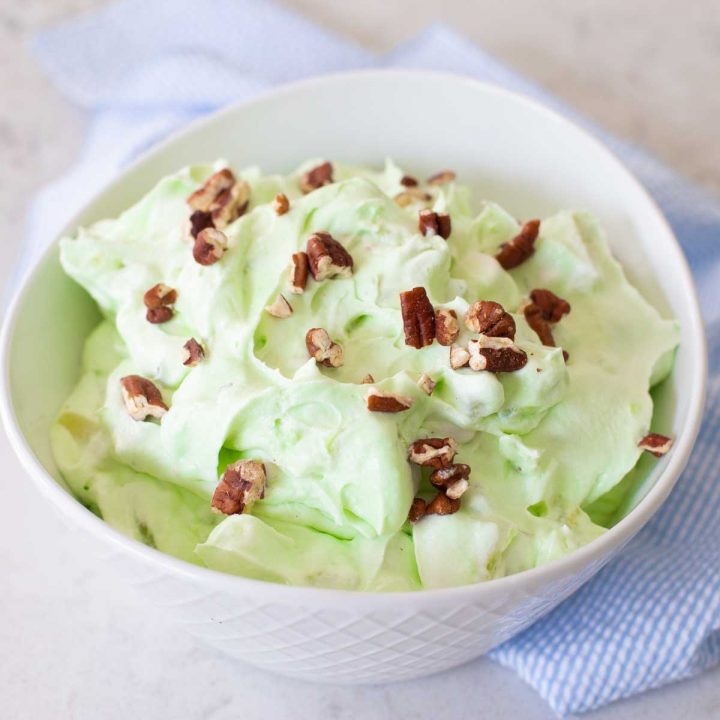 A large serving bowl of pistachio fluff has chopped pecans sprinkled over the top and a blue napkin on the side.