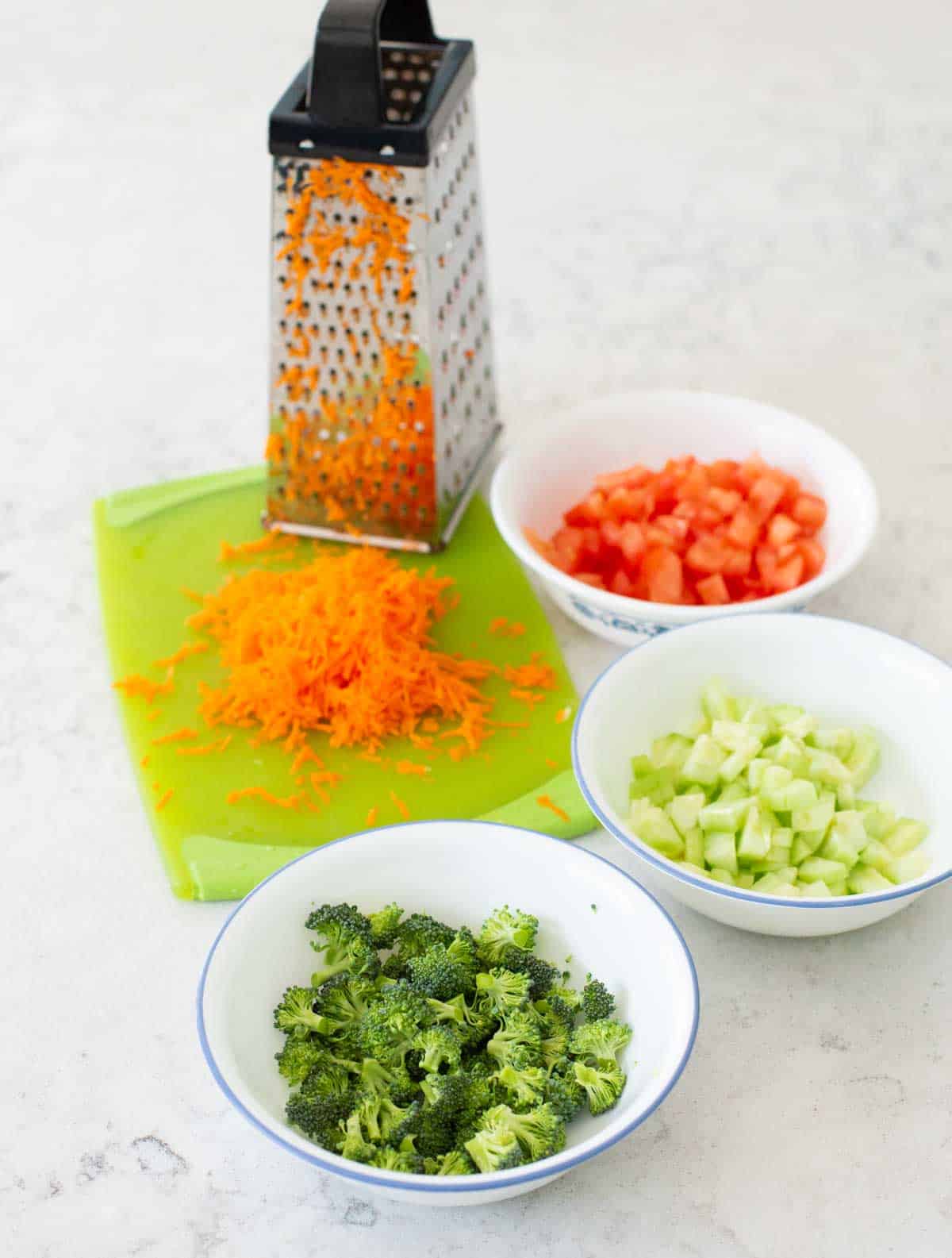 Bowls of raw broccoli, cucumber, tomato are on the counter with grated fresh carrot on a cutting board next to a box grater.