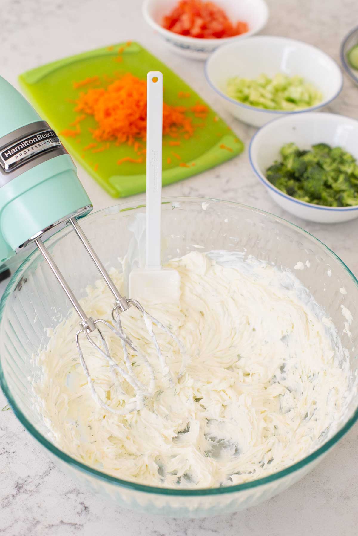 A mixing bowl has blended cream cheese with seasonings and prepped veggies are in the background.