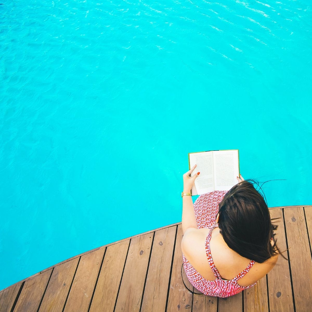 A woman reads her book at the edge of a big blue pool.