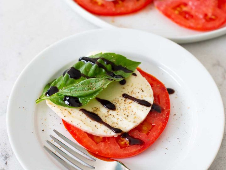 A sliced tomato, fresh mozzarella, fresh basil, and balsamic glaze on a plate with a fork.