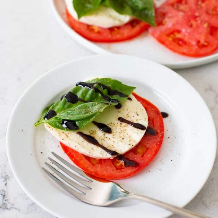 A sliced tomato, fresh mozzarella, fresh basil, and balsamic glaze on a plate with a fork.