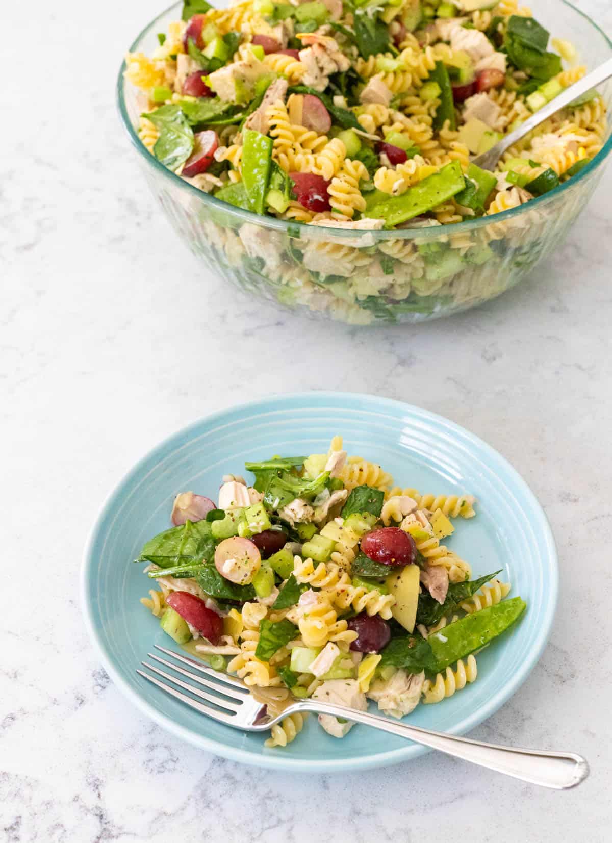 The serving bowl of pasta salad sits next to a blue plate filled with a portion and a fork.
