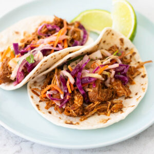 A spiced shredded chicken taco has slaw on the top with flour tortillas. Fresh lime wedges in the back.