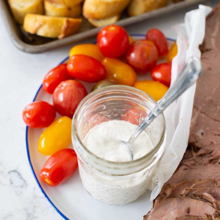 Homemade sour cream horseradish sauce in a mason jar on a plate next to roast beef and cherry tomatoes for dipping.