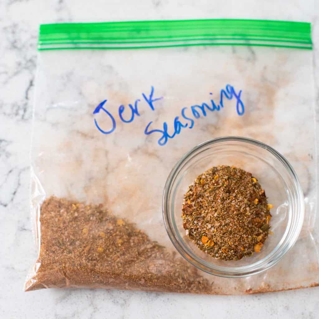 Homemade jerk seasoning blend is in a small glass seasoning bowl sitting on top of a plastic storage baggy labelled "Jerk Seasoning"