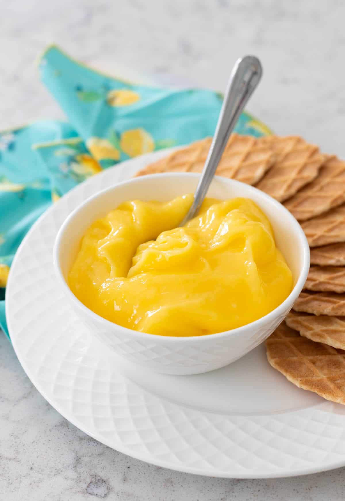 The bowl of bright yellow lemon curd has a spoon for serving. It rests on a plate of waffled butter cookies with slices of fresh lemon off to the side.