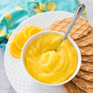 The bowl of bright yellow lemon curd has a spoon for serving. It rests on a plate of waffled butter cookies with slices of fresh lemon off to the side.