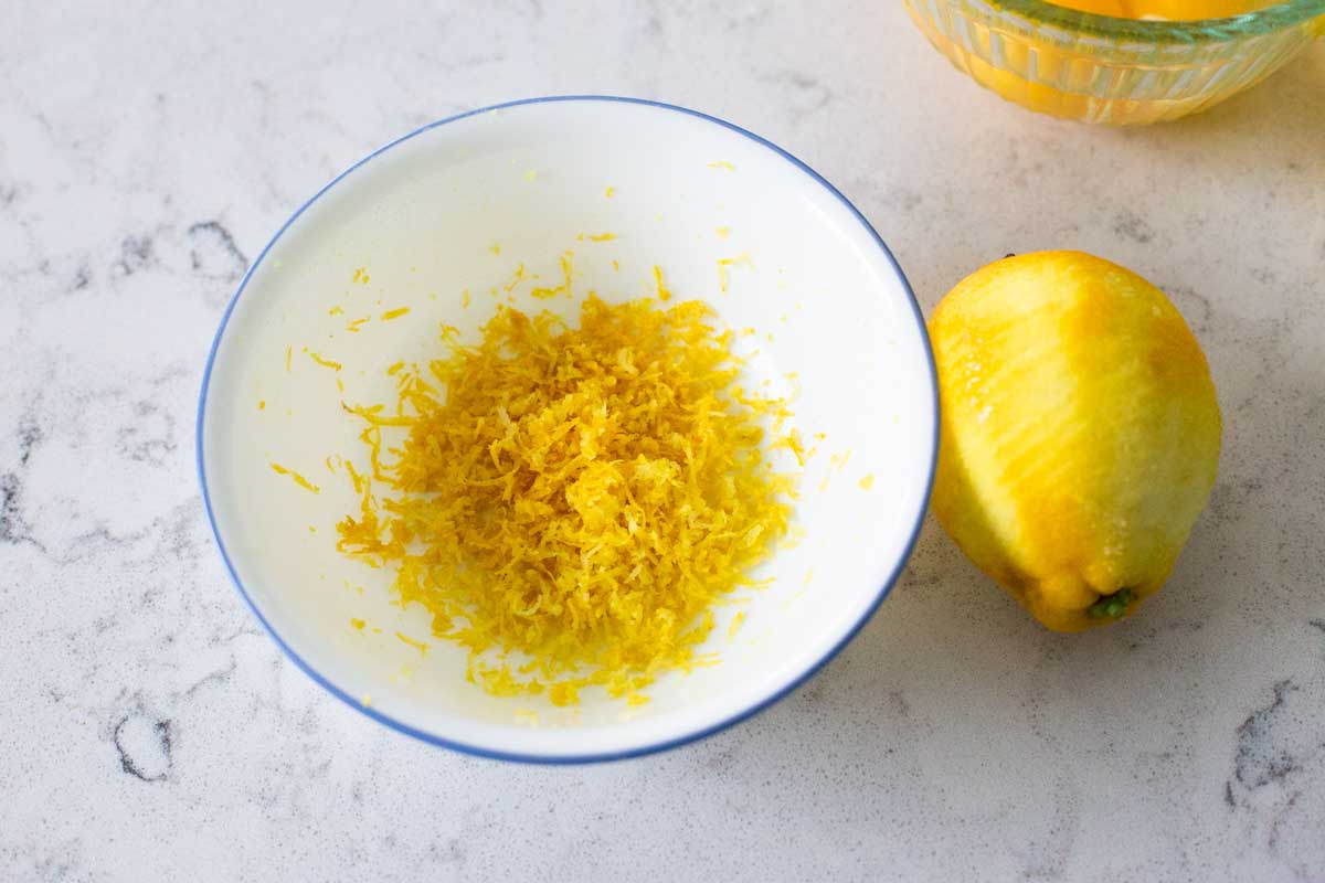A bowl of lemon zest sits next to a lemon that was used to get the zest.