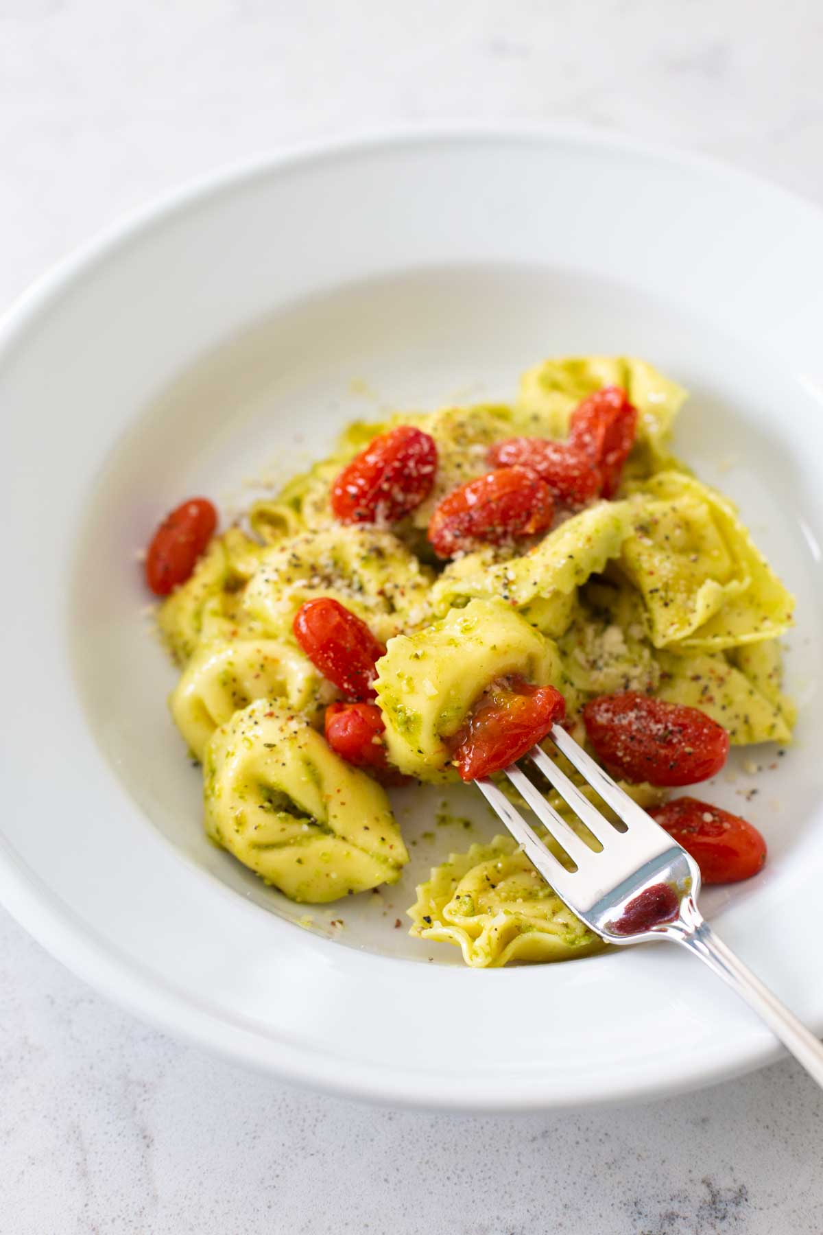 A serving of cheese tortellini in pesto sauce with roasted cherry tomatoes. There's a fork about to take a bite.