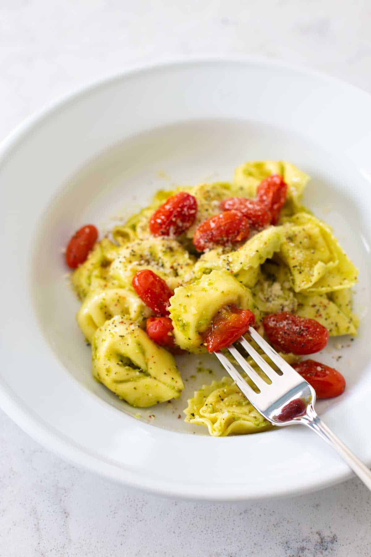 A serving of cheese tortellini in pesto sauce with roasted cherry tomatoes. There's a fork about to take a bite.