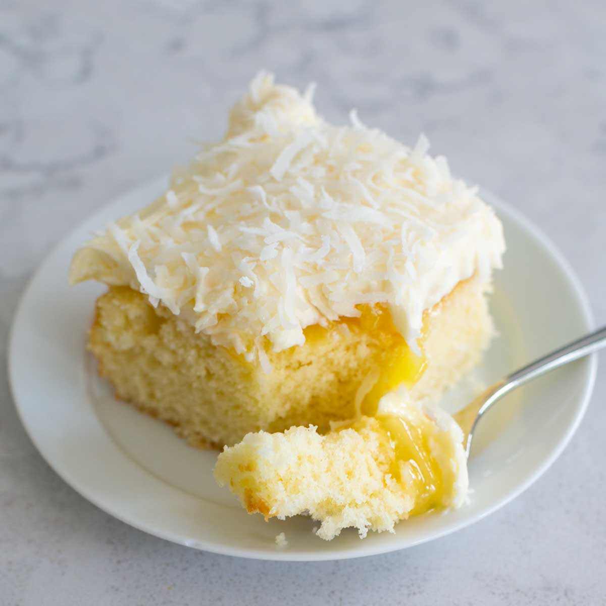 A slice of coconut cake with cream cheese frosting and shredded coconut has a layer of bright yellow pineapple filling.